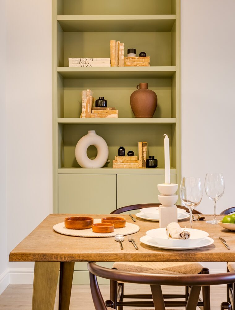 A wooden dining table decorated with a dining set and fruit bowl, alongside a green book shelf.