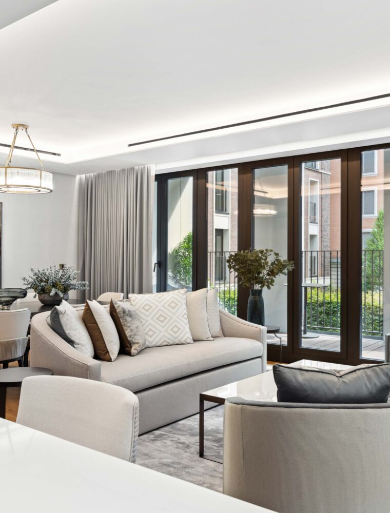 The lounge area of a London luxury apartment sourced by a London buyers agent. The lounge space features modern, grey furnishings, including a sofa and two chairs.